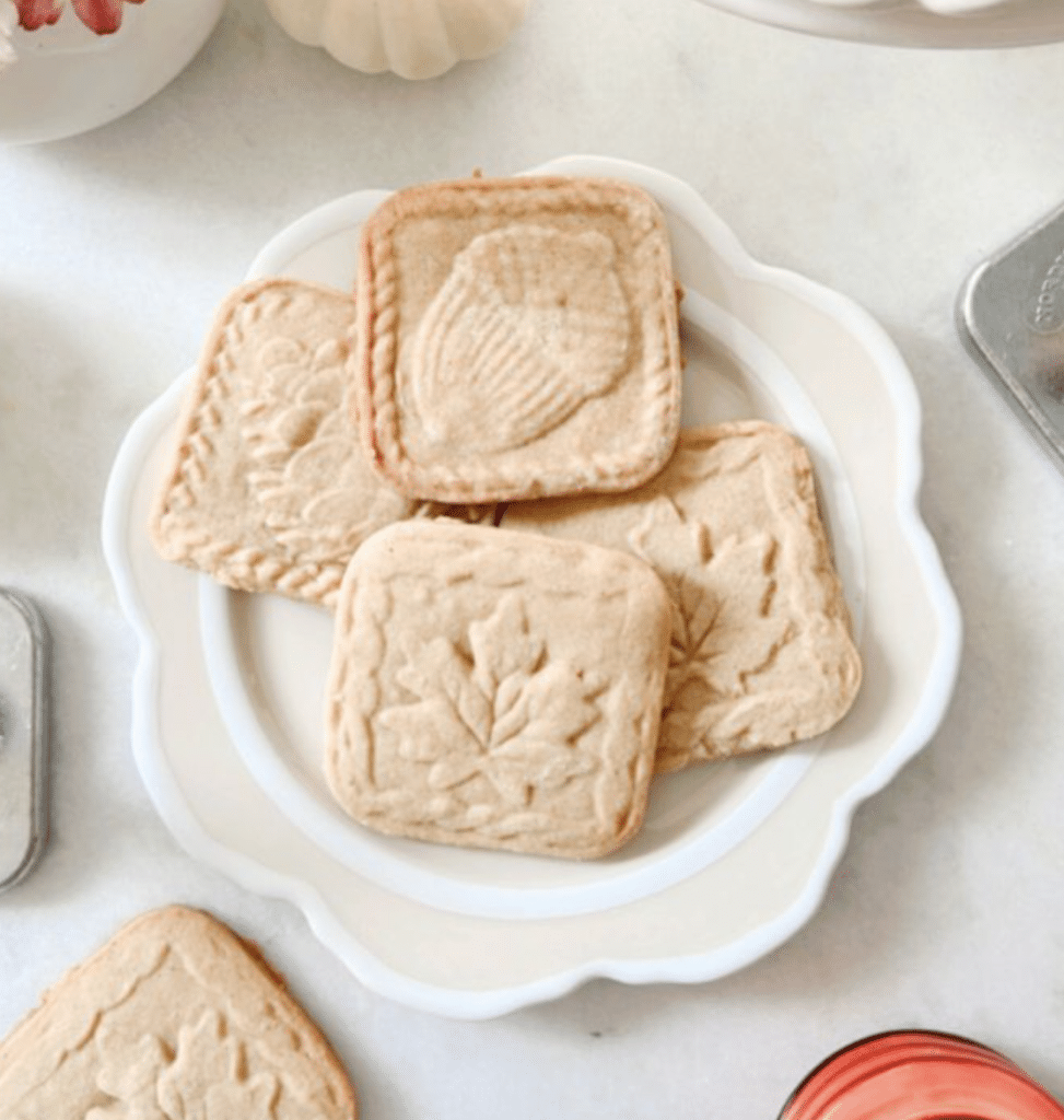 A plate of square, embossed cookies with various leaf and floral designs, perfect as Halloween treats for kids, sits on a white table. Additional cookies and a red glass are visible around the plate.